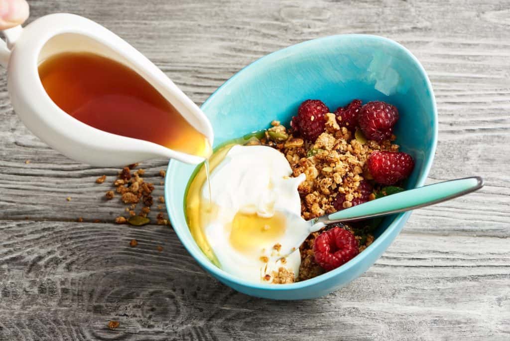 homemade granola in a bowl with raspberries and maple syrup on a wooden table.